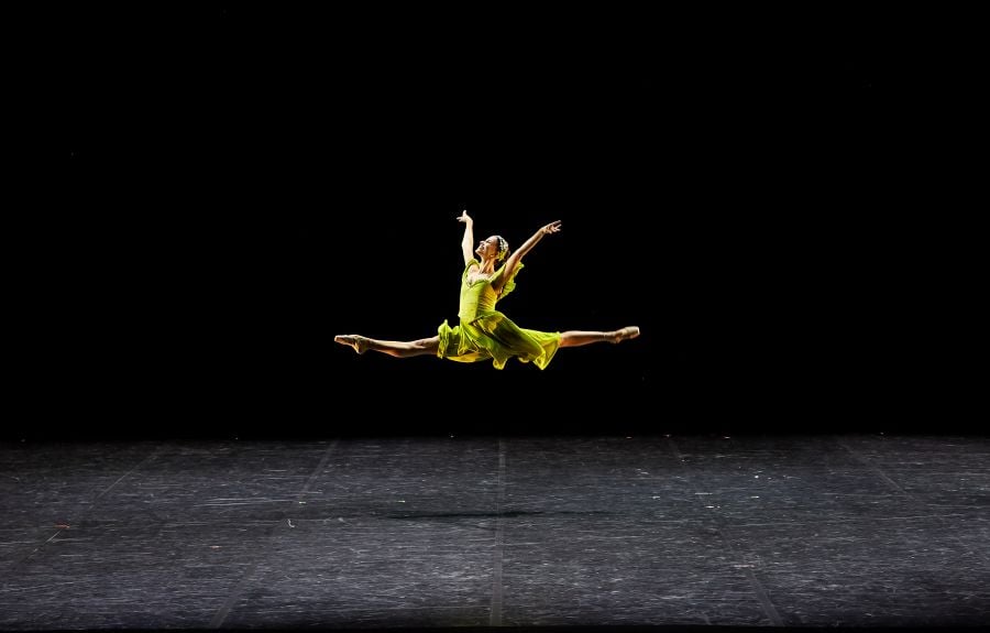 Georgia Waite, wearing a yellow dress, photographed executing a Grand Jeté in Chihiro Nomura’s Night Symphony Colours for Ballet at the Quarry: Incandescence. Her legs are spread a full 180° (grand écart) at the peak of the jump.