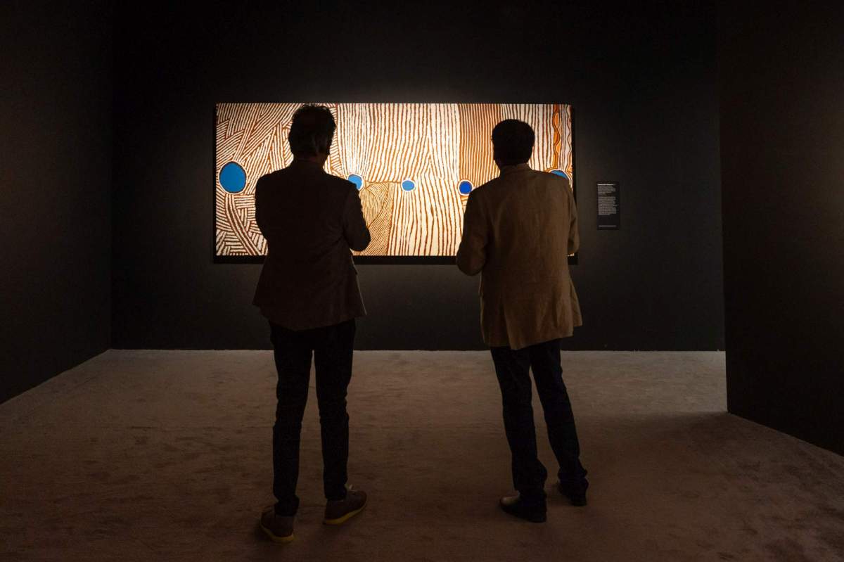 Songlines: Tracking the Seven Sisters: the silhouettes of two people standing in a darkened space looking at an Aboriginal dot painting shown spot-lit on a black gallery wall.