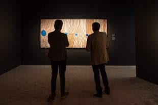 Songlines: Tracking the Seven Sisters: the silhouettes of two people standing in a darkened space looking at an Aboriginal dot painting shown spot-lit on a black gallery wall.