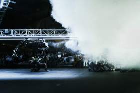 U>N>I>T>E>D Chunky Move, a dark stage with dramatic smoke effects and white lighting showing a female performer with robotic metallic costuming moving across the stage under a scaffolding set piece.