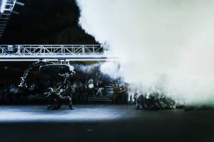 U>N>I>T>E>D Chunky Move, a dark stage with dramatic smoke effects and white lighting showing a female performer with robotic metallic costuming moving across the stage under a scaffolding set piece.