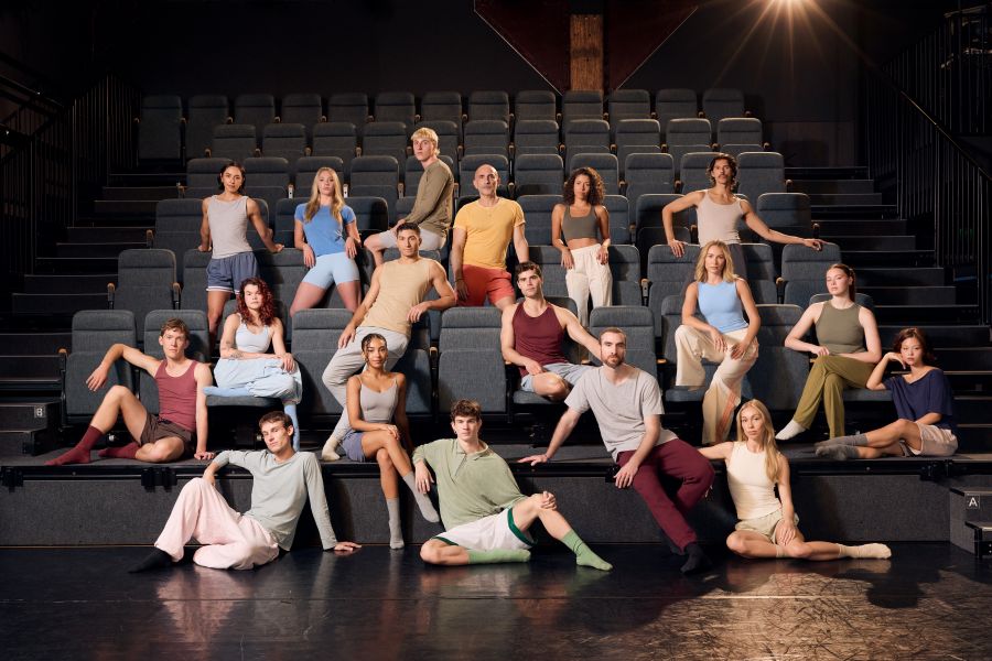 The Sydney Dance Company Ensemble and Artistic Director Rafael Bonachela. 18 people in loose clothes sit, lounge and stand in a seating bank in a theatre space.