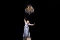 Kala Gare in the MTC production, My Brilliant Career. A young woman in a Victorian-era dresses stands atop a piano, holding one fist up triumphantly; an ornate chandelier hangs above her.