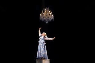 Kala Gare in the MTC production, My Brilliant Career. A young woman in a Victorian-era dresses stands atop a piano, holding one fist up triumphantly; an ornate chandelier hangs above her.