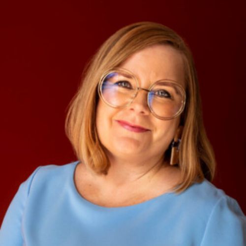 Natalie Jenkins is the new Performing Lines Board Chair. Jenkins is a middle-aged, fair-skinned woman with shoulder length, red-blonde hair. She wears large glasses and a blue blouse, tilts her hair a little to the left and smiles warmly at the camera.
