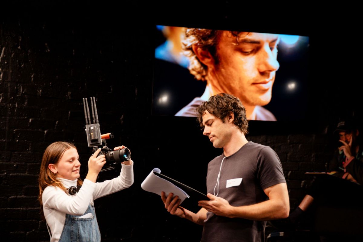 An earlier performance of POV, featuring different cast members than the performance reviews. n 11-year-old girl holds a sophisticated camera complete with antennae to broadcast it to screens in the theatre. She is filming a male actor, whose image is visible on a large screen behind them.