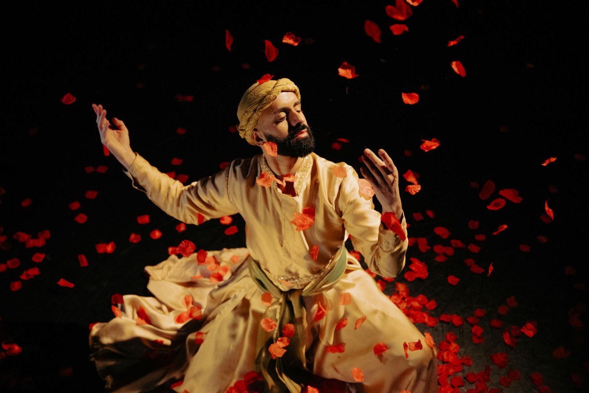 Songs of the Bulbul Aakash Odedra: a young male dancer in a white turban and robe photographed spinning with his eyes closed under a shower of red rose petals that are descending on him from above.