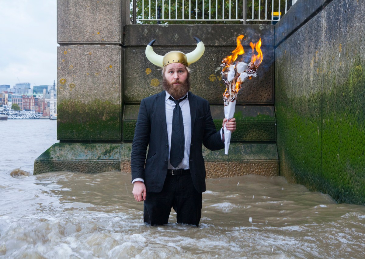 James Rowland: Team Viking. A fair-skinned man with straw-blonde hair and a reddish beard stands thigh deep in the waters of the Thames. He is wearing a plastic Viking helmet with ahistorical horns, a black suit, white business shirt and black tie, and carries a burning torch made of rolled-up paper.