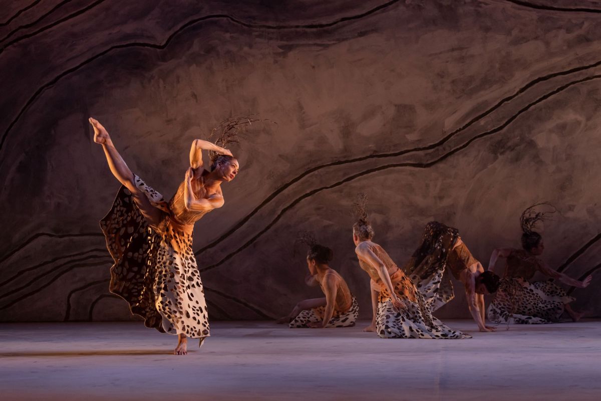 Bangarra's Terrain at Sydney Opera House. The photo shows an Aboriginal woman dancing, caught mid-routine, with her right leg dramatically raised; her right arm is bent above her head. Other dancers are visible, kneeling on the stage behind her.