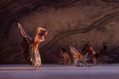 Bangarra's Terrain at Sydney Opera House. The photo shows an Aboriginal woman dancing, caught mid-routine, with her right leg dramatically raised; her right arm is bent above her head. Other dancers are visible, kneeling on the stage behind her.