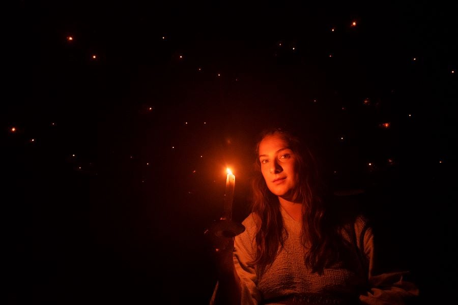 A woman lit only by the candle she holds in her right hand gazes at the camera, a sea of stars behind her. A scene from The Childhood of the World, part of Slingsby’s A Concise Compendium of Wonder at Adelaide Festival 2026. Photo: Eyefood.