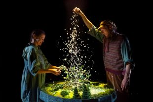 Two actors stand over a miniature walled garden. One actor, bearded and wearing a cap, lets a handful of stage snow rain down as the other actor, who is female-presenting, watches on delightedly. A scene from The Giant's Garden, part of Slingsby's A Concise Compendium of Wonder at Adelaide Festival 2026.