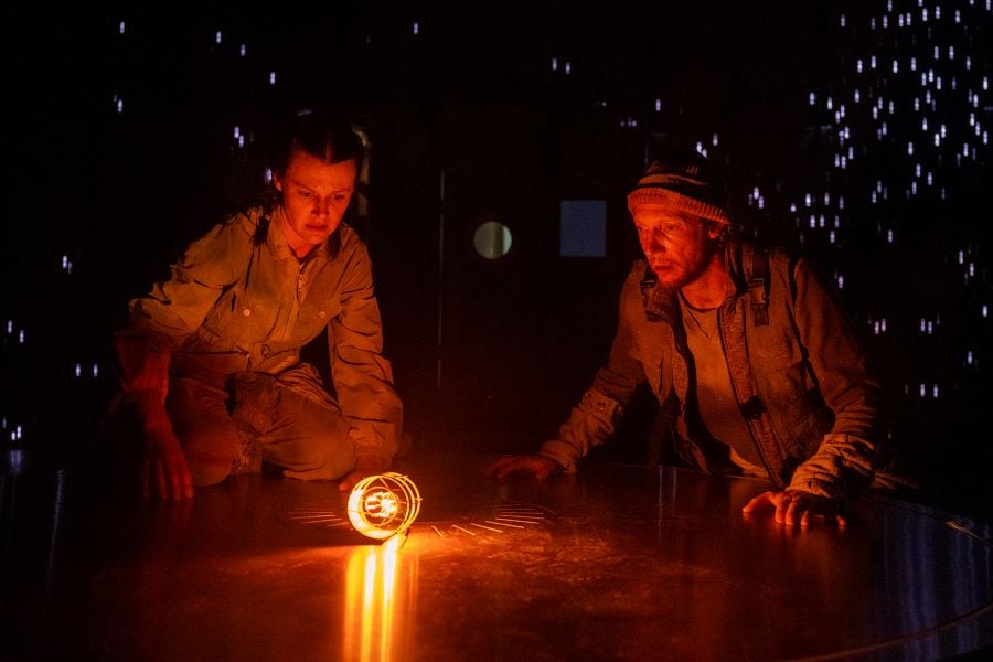 A scene from Slingsby's The Tree of Light, part of A Concise Compendium of Wonder at Adelaide Festival 2026. Two actors crouch around a low, circular structure, gazing at and illuminated by a golden light; blurred stage stars shine behind them.