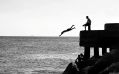 A black and white photograph of a person diving off a pier as another person sits watching. arts sector appointments