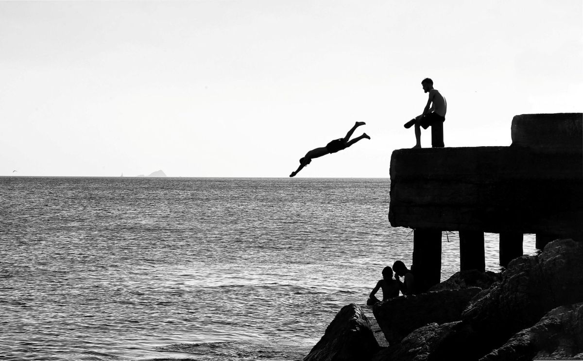 A black and white photograph of a person diving off a pier as another person sits watching. arts sector appointments