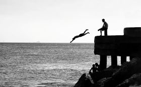 A black and white photograph of a person diving off a pier as another person sits watching. arts sector appointments