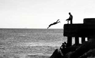 A black and white photograph of a person diving off a pier as another person sits watching. arts sector appointments