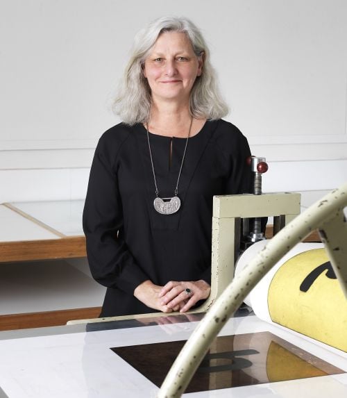 Anne Virgo OAM, Artistic Director and CEO of Australian Print Workshop. A grey-haired woman dressed in black, and wearing a silver necklace, stands in front of a printing press. defunded victorian arts organisations