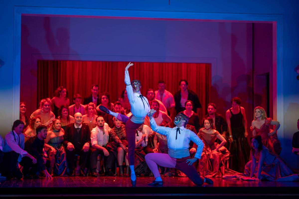 La traviata West Australian Opera: a photograph of an opera performance showing two Spansih matador style masked dancers posing in front of a opera chorus ensemble in Victorian-era costume on a stage.