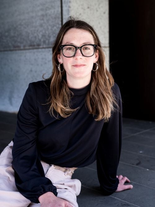 Harriet Devlin, Arena Theatre Company's new Creative Producer. A fair-skinned woman with long brown hair and wearing dark-framed glasses, sits and smiles at the camera.