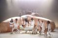 A scene from Florentina Holzinger's 'A year without summer', programmed for RISING 2026. Ten culturally and physically diverse performers wearing hospital whites (including a person lying on a hospital bed and another using a walking frame) sing in front of a larger-than-life nude female torso. Six more white-clad performers are visible at the rear of the stage, performing on a raised, industrial-looking platform.