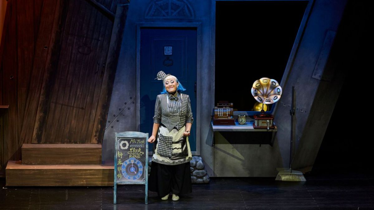 CDP Theatre Producers: Klokwerk production in China. A photo of a young female Asian performer standing on stage dressed in a black and white checkered costume surrounded by some old fashioned set pieces such as a gramophone and an old cash register machine.