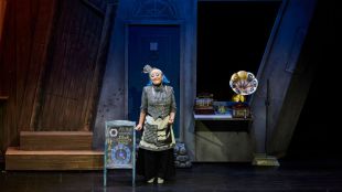 CDP Theatre Producers: Klokwerk production in China. A photo of a young female Asian performer standing on stage dressed in a black and white checkered costume surrounded by some old fashioned set pieces such as a gramophone and an old cash register machine.