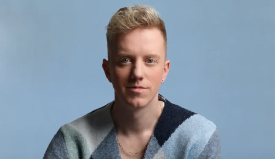 James Barr's Sorry I Hurt Your Son (Said My Ex to My Mum) at Melbourne International Comedy Festival. A blonde-haired, fair-skinned man wearing a blue and white patterned jacket over a white t-shirt. He is smiling softly at the camera.