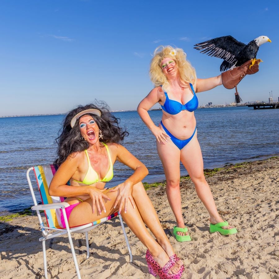 Alex Hines & Sarah Stafford's Birds at Melbourne International Comedy Festival 2026. Two women in swimwear and over the top make-up and wigs pose at the beach. One of them has a bald eagle toy perched on her leather-gloved wrist like a hunting falcon.