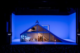 Simon Stone's The Cherry Orchard opened Adelaide Festival 2026. Actors stand in front of a glass-fronted house on a large stage, lit blue to indicate it is nighttime. Audience members heads are visible; the photo has been taken in the auditorium, some distance from the stage.