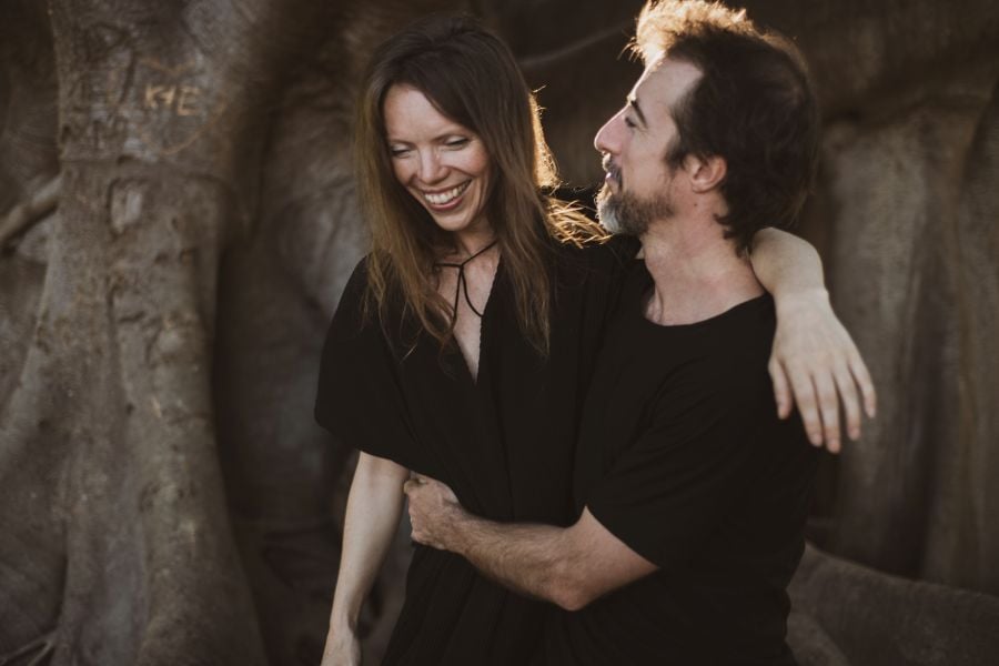 L-R: Dancenorth Australia's co-Artistic Directors Amber Haines and Kyle Page. Both are fair-skinned and wearing black; they're posed in a warm embrace in front of a tree.  Amber has long brown hair and is looking down, towards the ground; Kylehas a short brown beard tinged with grey and short black hair.