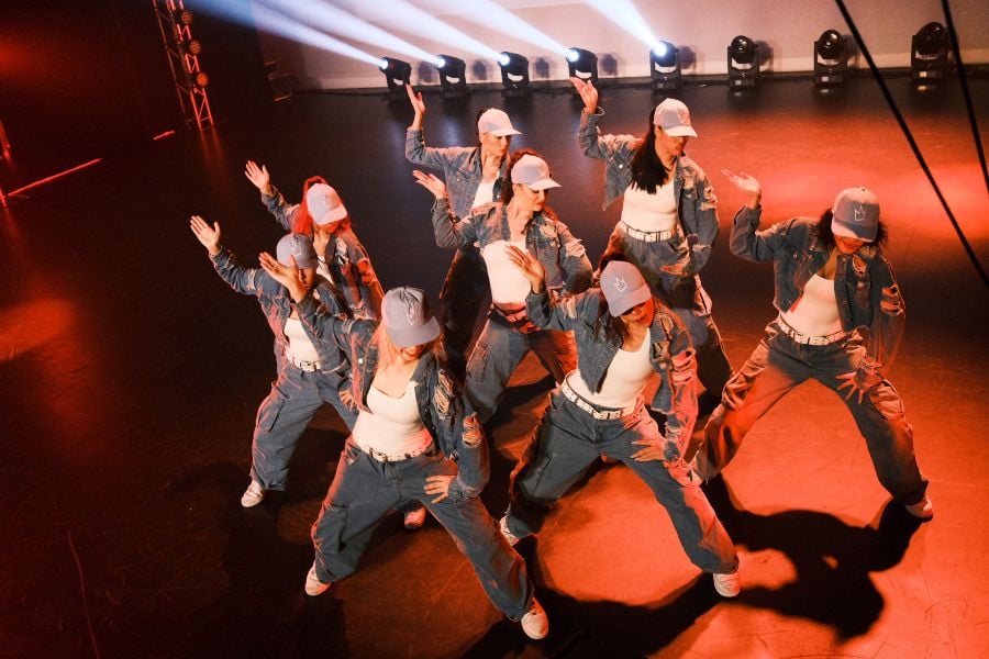 Eight female-presenting members of the Royal Family Dance Crew perform Defend The Throne, which has been programmed for RISING 2026. The dancers all wear white sneakers, white t-shirts, green pants and jackets and grey baseball caps. All eight women stand in the same pose, with their right hands raised and their left hands resting at the top of their left leg.