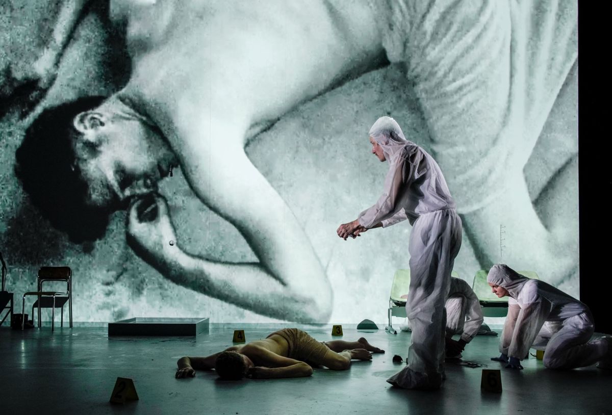 History of Violence at Adelaide Festival 2026. A half-naked actor, covered by a bedsheet, sprawls on stage. His image is cast to a screen behind him by the camera held in a second actor's hands; the second actor wears a white plastic forensic jumpsuit.