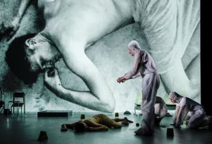 History of Violence at Adelaide Festival 2026. A half-naked actor, covered by a bedsheet, sprawls on stage. His image is cast to a screen behind him by the camera held in a second actor's hands; the second actor wears a white plastic forensic jumpsuit.