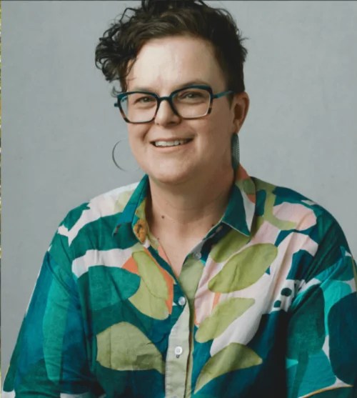 Justine Hyde, Abbotsford Convent Foundation CEO. A smiling, fair skinned woman with artfully tousled dark brown hair. She wears glasses, large circular earings and a green, white and orange patterned blouse.