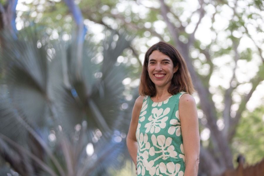 Darwin Festival's outgoing Artistic Director Kate Fell. A fair-skinned woman in a sleeveless green and white patterned dress; she has shoulder length brown hair and smiles at the camera. Tropical plants and trees can be seen behind her though they are a little out of focus.