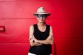 The late Rhoda Roberts AO. The Bundjalung Widjabul Wiyebal woman is pictured standing in front of a red-painted wall with her arms folded; she wears a black t-shirt, a broad-brimmed hat and dark-framed glasses.