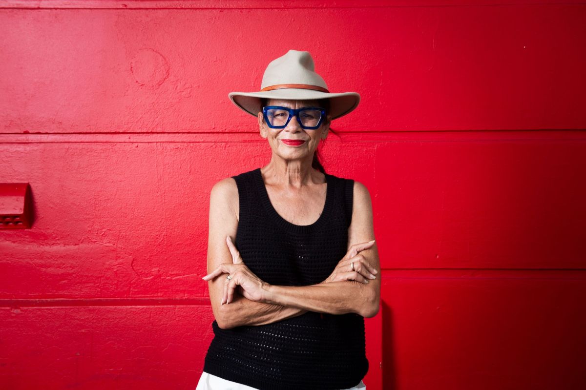 The late Rhoda Roberts AO. The Bundjalung Widjabul Wiyebal woman is pictured standing in front of a red-painted wall with her arms folded; she wears a black t-shirt, a broad-brimmed hat and dark-framed glasses.
