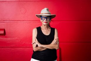 The late Rhoda Roberts AO. The Bundjalung Widjabul Wiyebal woman is pictured standing in front of a red-painted wall with her arms folded; she wears a black t-shirt, a broad-brimmed hat and dark-framed glasses.