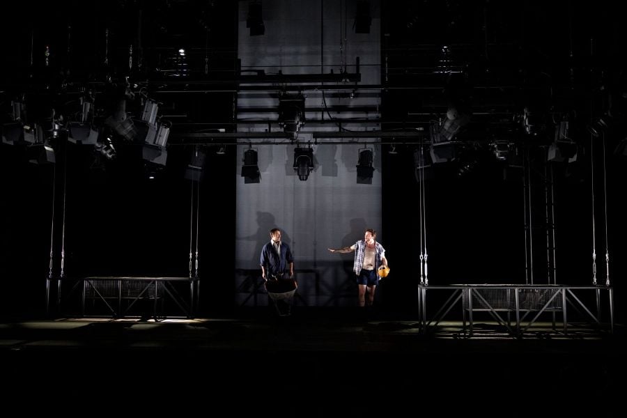 Steve Bastoni and Darcy Kent on the set of MTC's 2026 production, West Gate. The two actors, playing builders, stand before a vast slab of 'concrete' representing the under-construction West Gate Bridge. The lighting rig has been lowered to stage level to emphasise the industrial nature of the set.