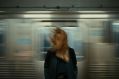 A woman stands on a train platform, her hair blowing around her face as a train speeds past behind her, blurred in movement. arts sector appointments