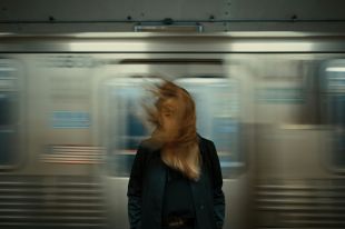 A woman stands on a train platform, her hair blowing around her face as a train speeds past behind her, blurred in movement. arts sector appointments
