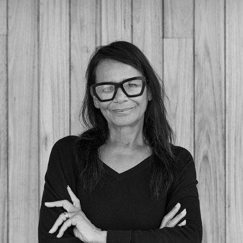 The late Rhoda Roberts AO. A black and white photo of Roberts, an Aboriginal woman; she has long black hair and wears a long sleeved black shirt and dark framed glasses, and stands with her arms crossed, smiling at the camera.