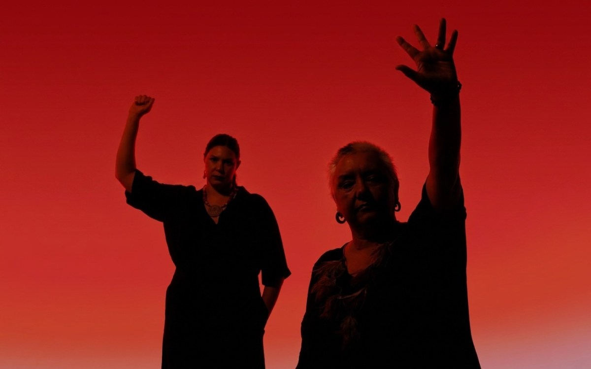 L-R: Allara and Dr Lou Bennett AM, the lead creatives of GANBINAN!, a 2027 YIRRAMBOI commission. The two Aboriginal women, of different generations, are posted in semi-silhouette against a red backdrop; Allara has her right fist raised in a Black power salute while Bennett has her left hand raised as if waving, her fingers spread.