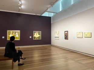 John Brack x Noel McKenna exhibition at the National Portrait Gallery: a woman dressed in black is sitting on a gallery bench looking at a range of small paintings hung on the gallery walls . One wall is a deep purple colour, the other is white.