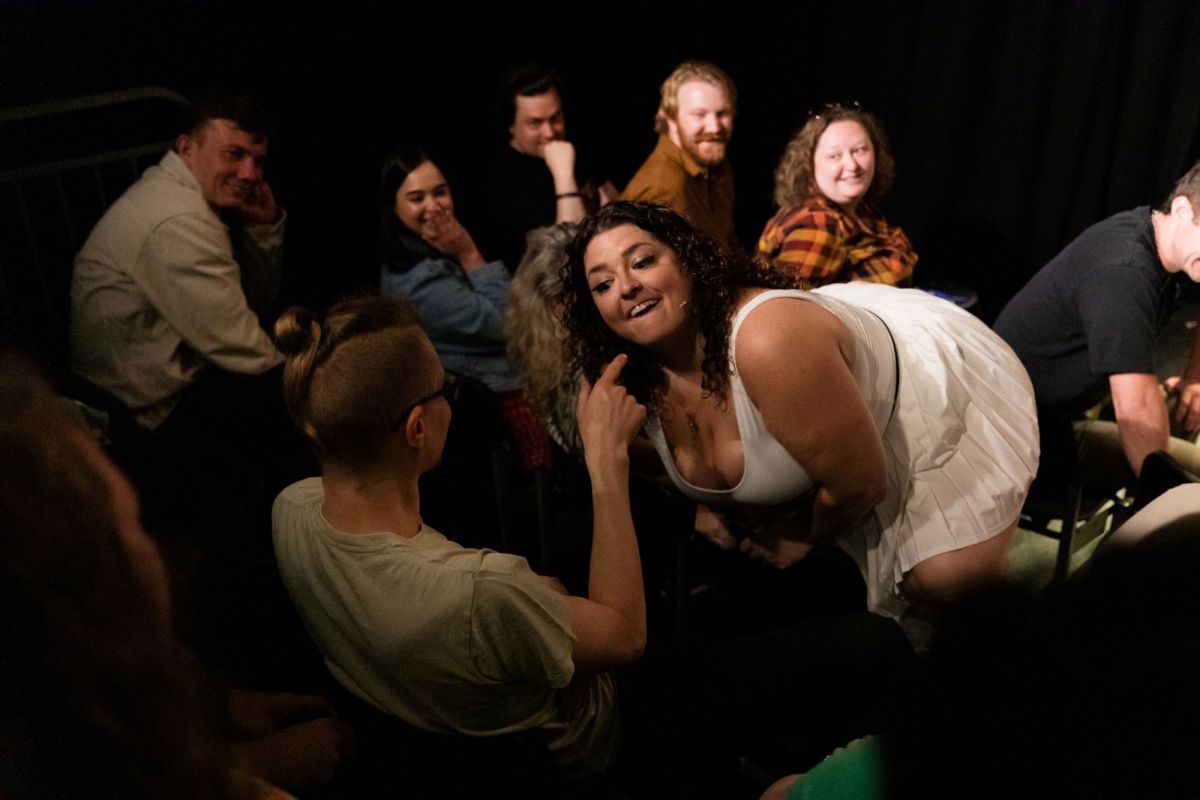 Comedian Mel McGlensey 's MICF show Mel McGlensey is Normal is one of many shows reviewed by writers for the Funny Tonne. The photo shows McGlensey, a curvacious figure with dark curly hair and wearing a sleeveless white dress, crouched in front of an audience member as if she is about to bit his raised finger. More audience members watch on in the background.