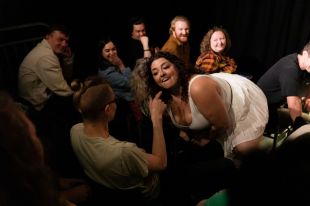 Comedian Mel McGlensey 's MICF show Mel McGlensey is Normal is one of many shows reviewed by writers for the Funny Tonne. The photo shows McGlensey, a curvacious figure with dark curly hair and wearing a sleeveless white dress, crouched in front of an audience member as if she is about to bit his raised finger. More audience members watch on in the background.