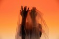 Jedda Atkinson-Costa, the lead creative of Withewa, one of the 2027 YIRRAMBOI commissions. A half-seen figure stands beneath a transparent veil, with their hands held up and fingers spread. They are photographed against a vivid orange background.