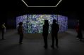 Khaled Sabsabi, conference of one's self, 2026, Australia Pavilion. Six people stand in a large, dark space, contemplating the installed artwork, which consists of a series of eight very large paintings placed together in an octagonal pattern; we can see three of the purple and green paintings, which are brightly lit by projections cast onto their surfaces.