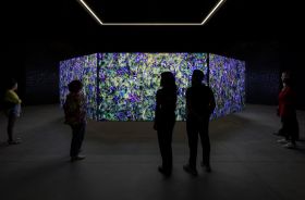 Khaled Sabsabi, conference of one's self, 2026, Australia Pavilion. Six people stand in a large, dark space, contemplating the installed artwork, which consists of a series of eight very large paintings placed together in an octagonal pattern; we can see three of the purple and green paintings, which are brightly lit by projections cast onto their surfaces.